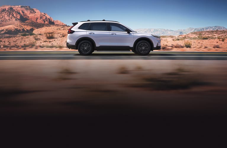 2027 Honda CR-V Sport Touring Hybrid in Platinum White Pearl profile driving on a scenic desert road (Compact Crossover SUV).